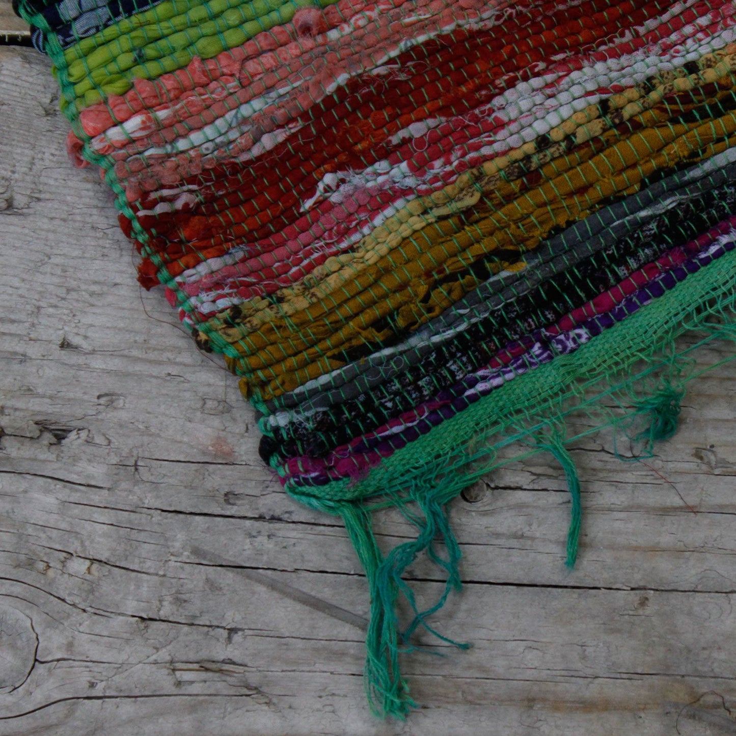 Corner detail of an Indian rag rug showing green fringe and textured weave on a rustic wood surface.