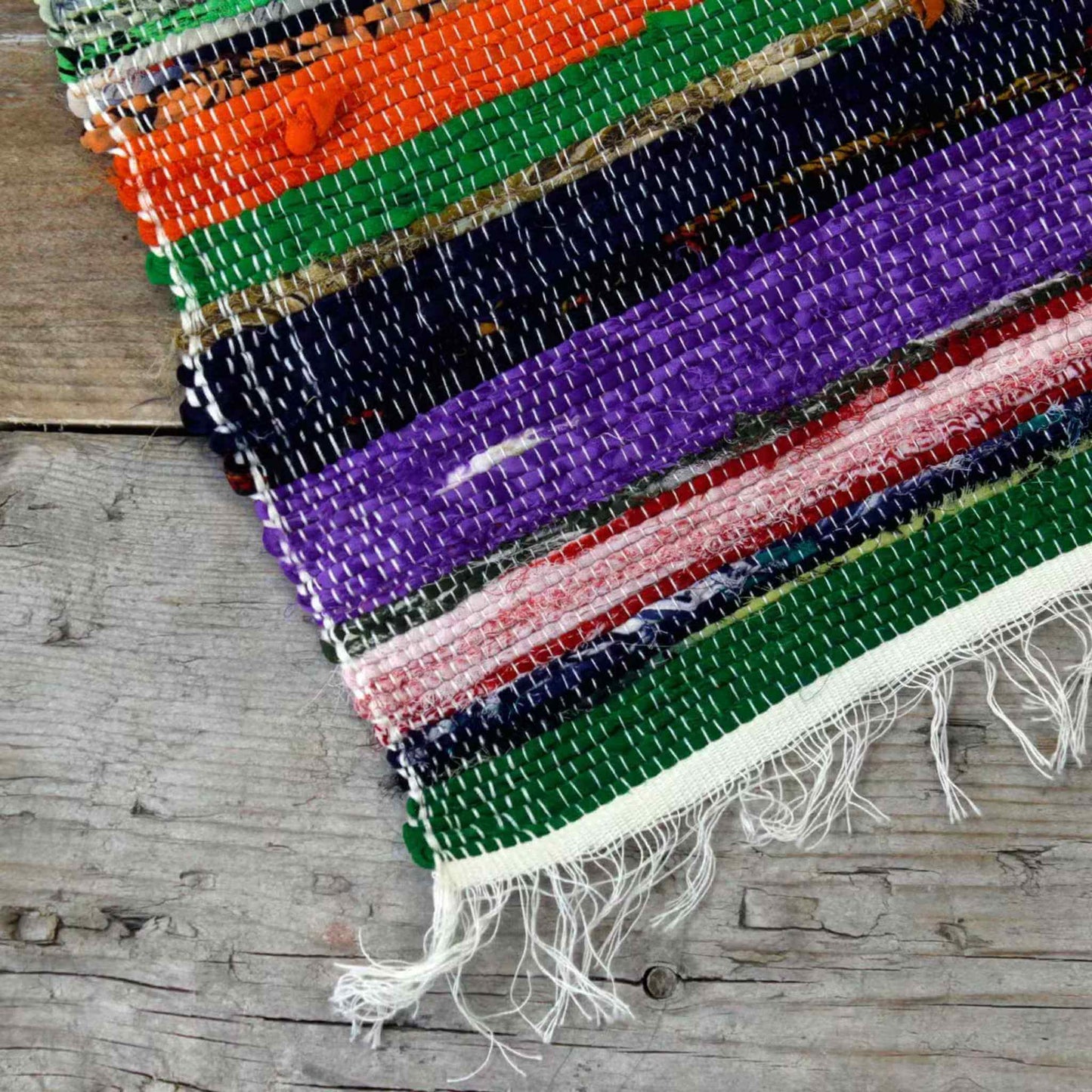 Corner of a multicoloured Indian rag rug with purple, green, orange, and black stripes on a wooden floor.