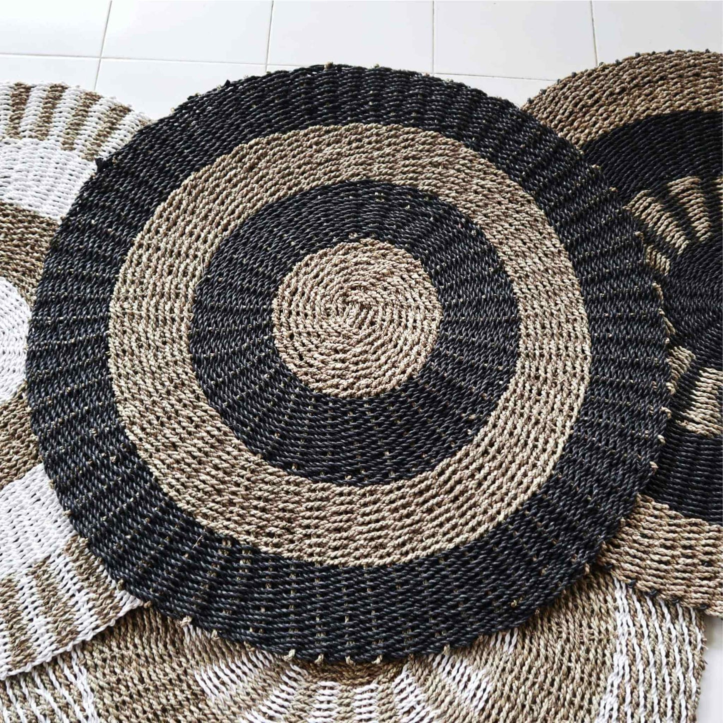 Round black and tan seagrass rug, 1m diameter, handwoven circular design, styled next to other natural rugs.