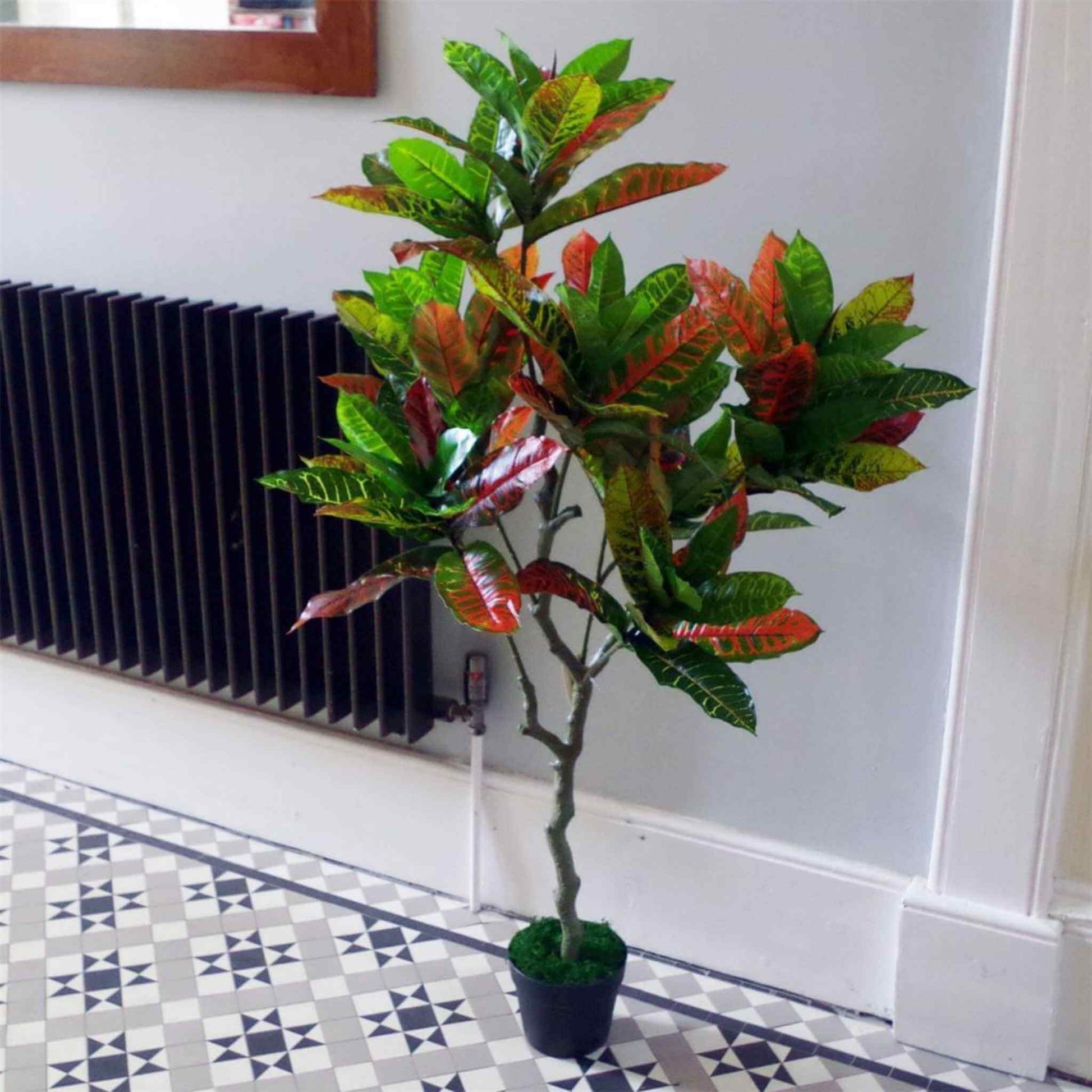 130cm artificial Codiaeum multicoloured tree with green, orange and yellow leaves in a black pot, placed in a hallway with a tiled floor and white walls.