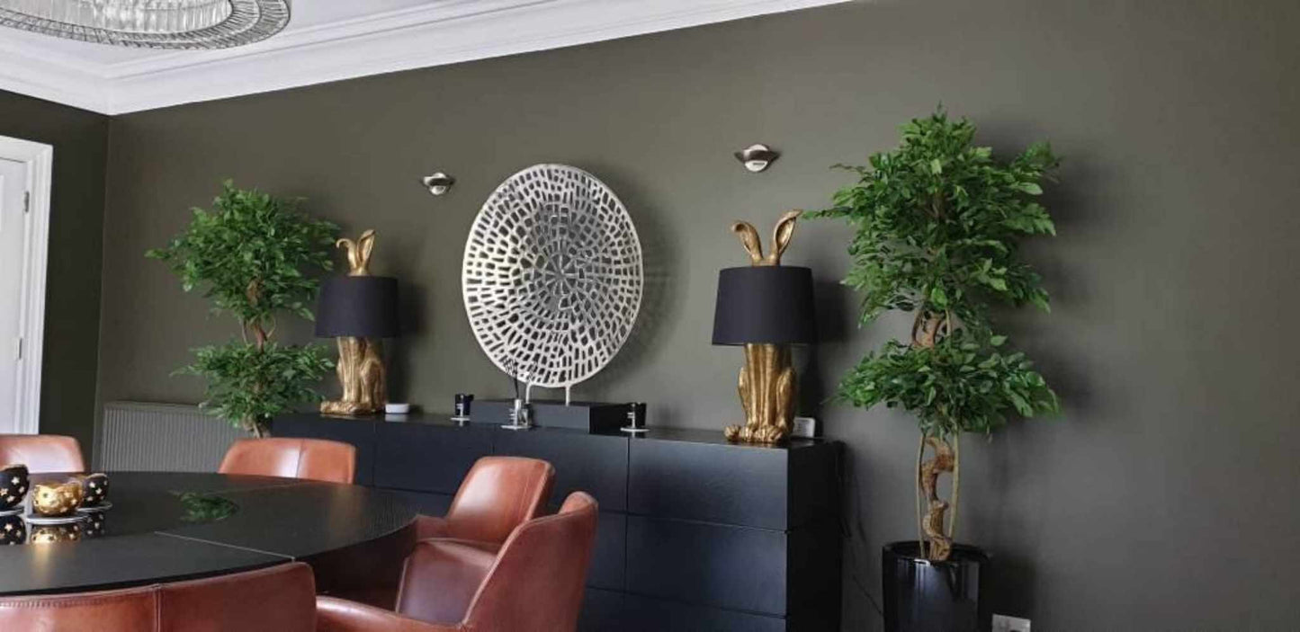 Artificial twisted ficus trees used symmetrically in a calm, contemporary dining room with dark green walls and gold accent lighting.