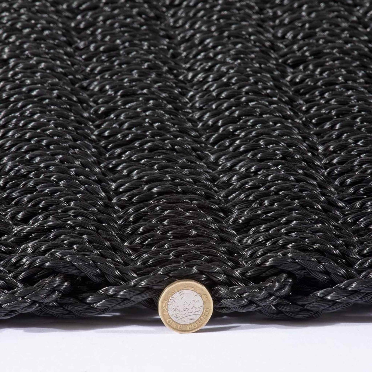 Side angle of braided polypropylene doormat with pound coin for scale, showing thickness and texture.