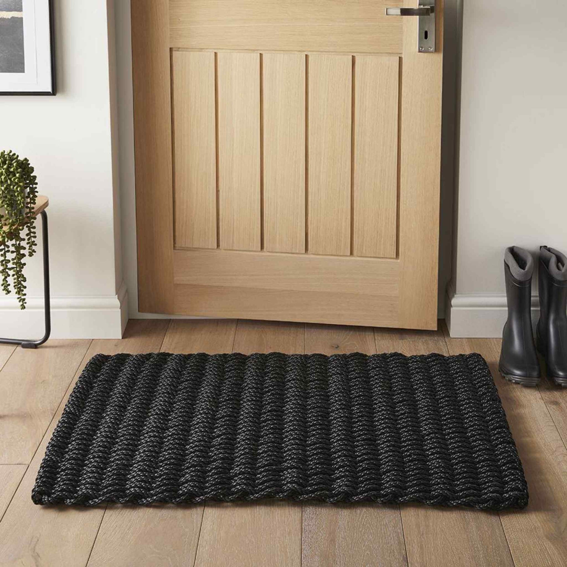 Charcoal braided doormat styled at a doorway on a wooden floor beside boots and a potted plant.