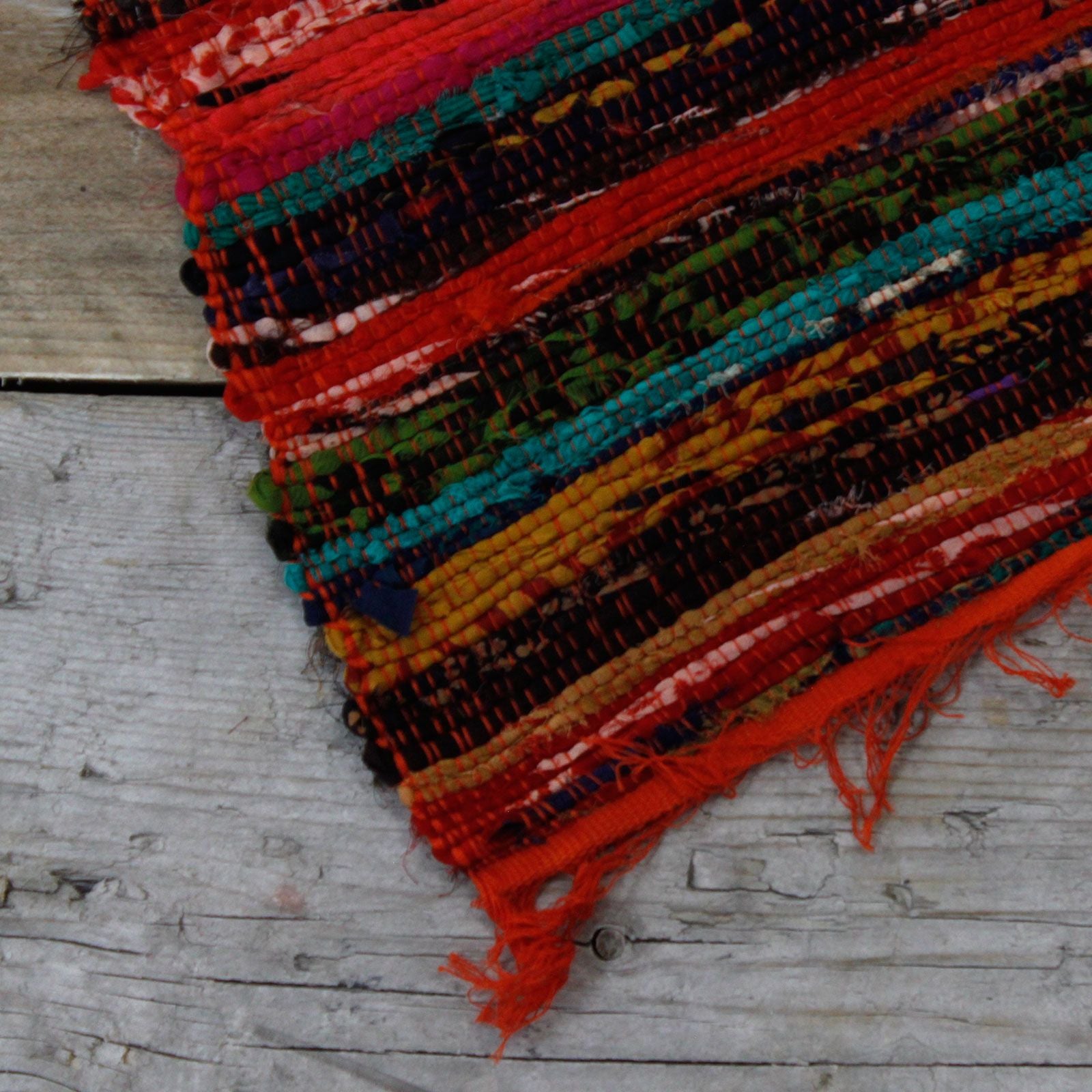 A woven orange cotton rag rug laid diagonally, showing intricate mix-fabric stripes and frayed edges.