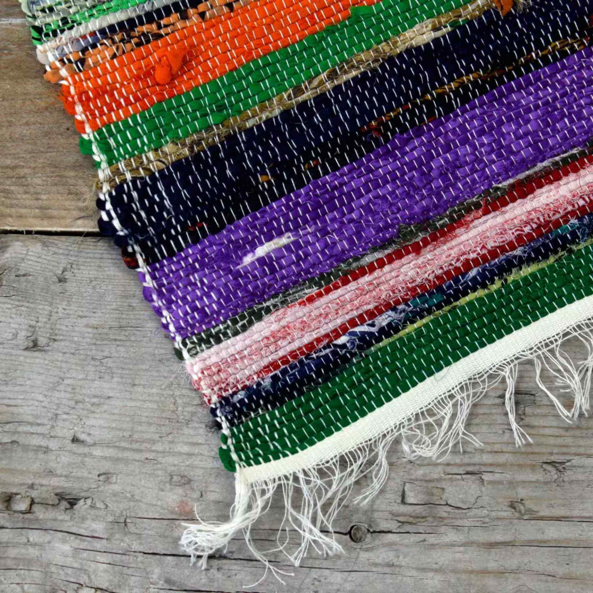 Corner of a multicoloured Indian rag rug with purple, green, orange, and black stripes on a wooden floor.