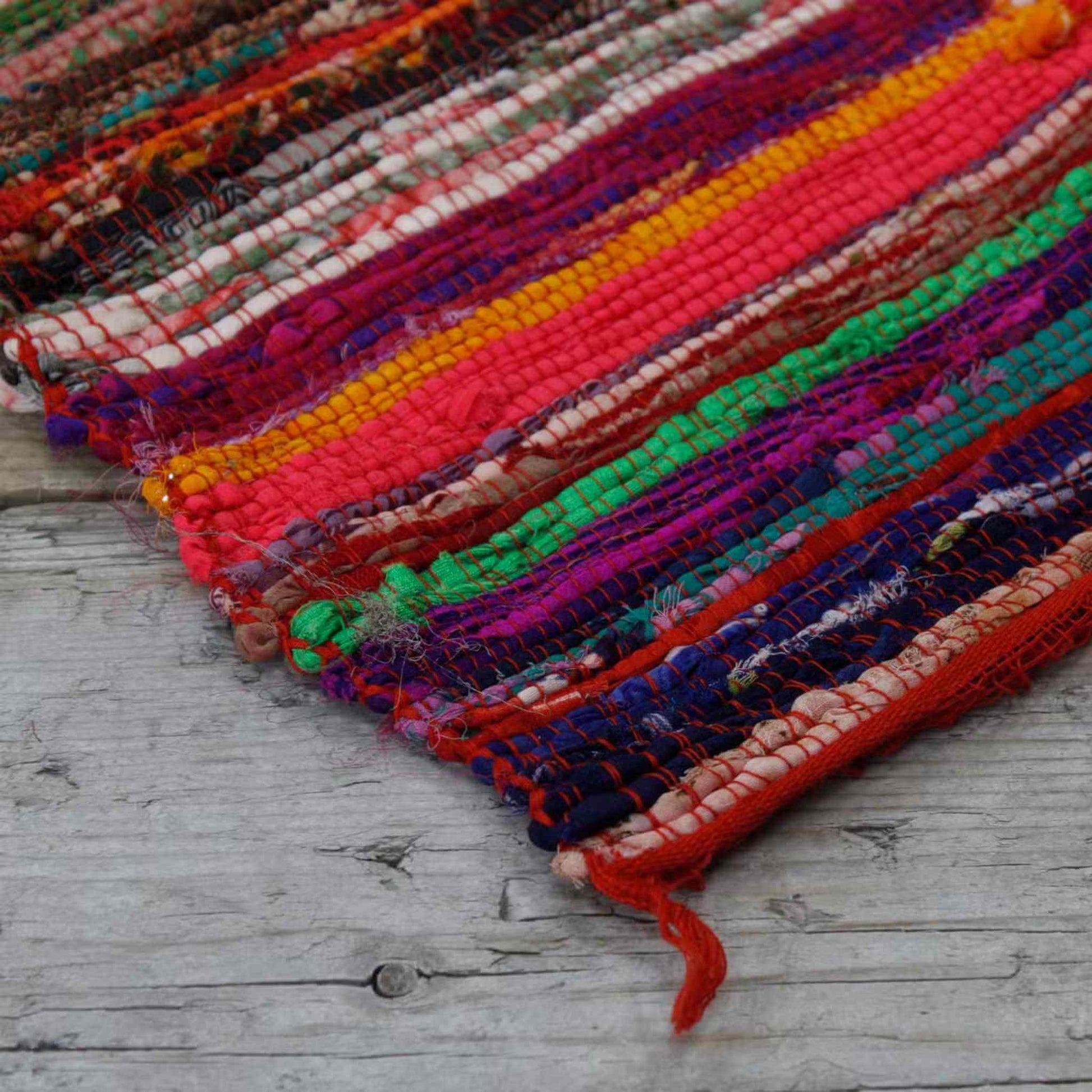 Close-up of the corner of a red striped rag rug revealing bright woven threads in green, pink, and blue on a wooden floor.