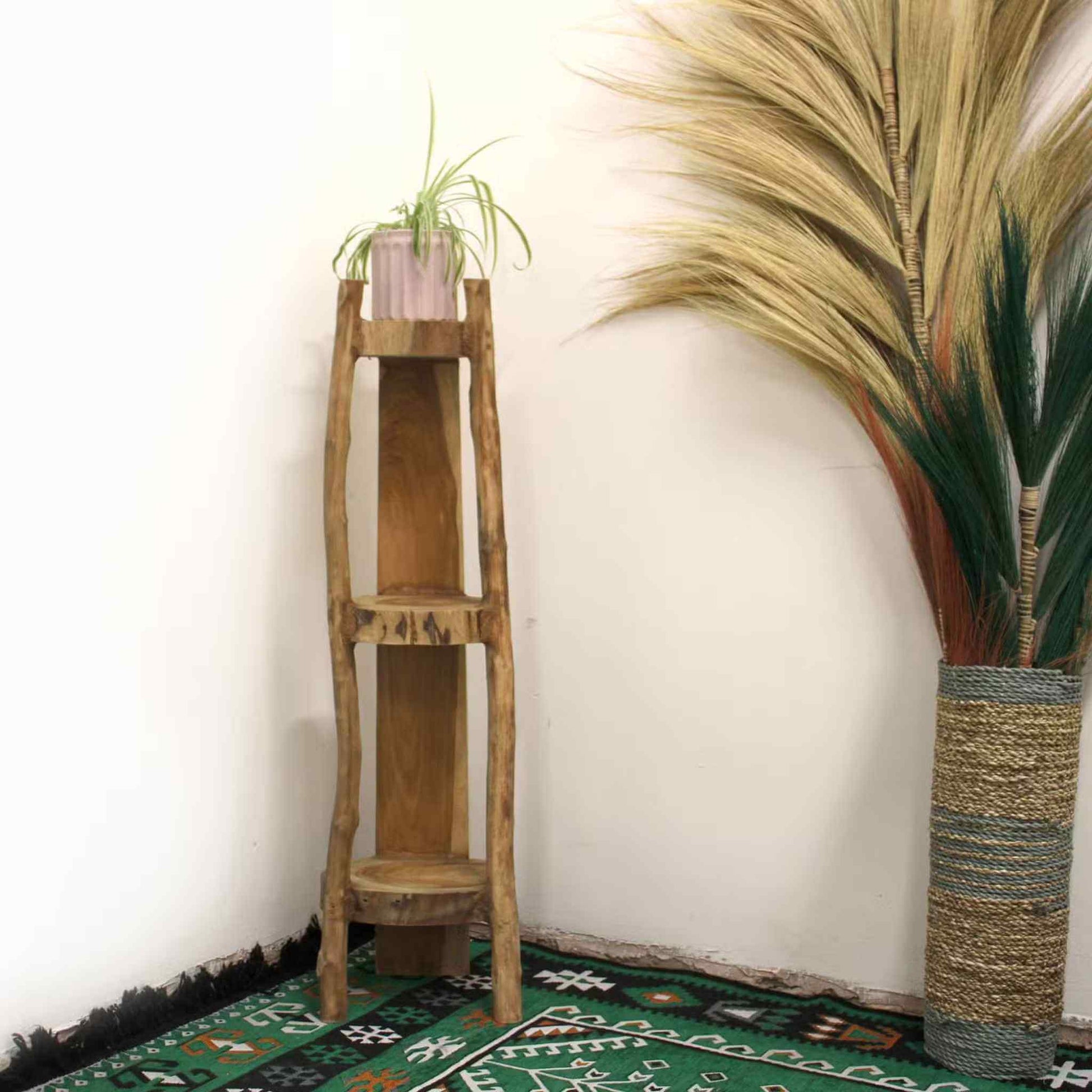 Natural teak corner shelf displayed in a living area corner with a green patterned rug, next to a tall vase of dried grass.