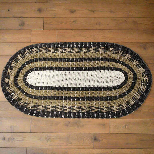 Overhead view of oval seagrass rug in neutral tones of black, tan, and white, set on wooden flooring.