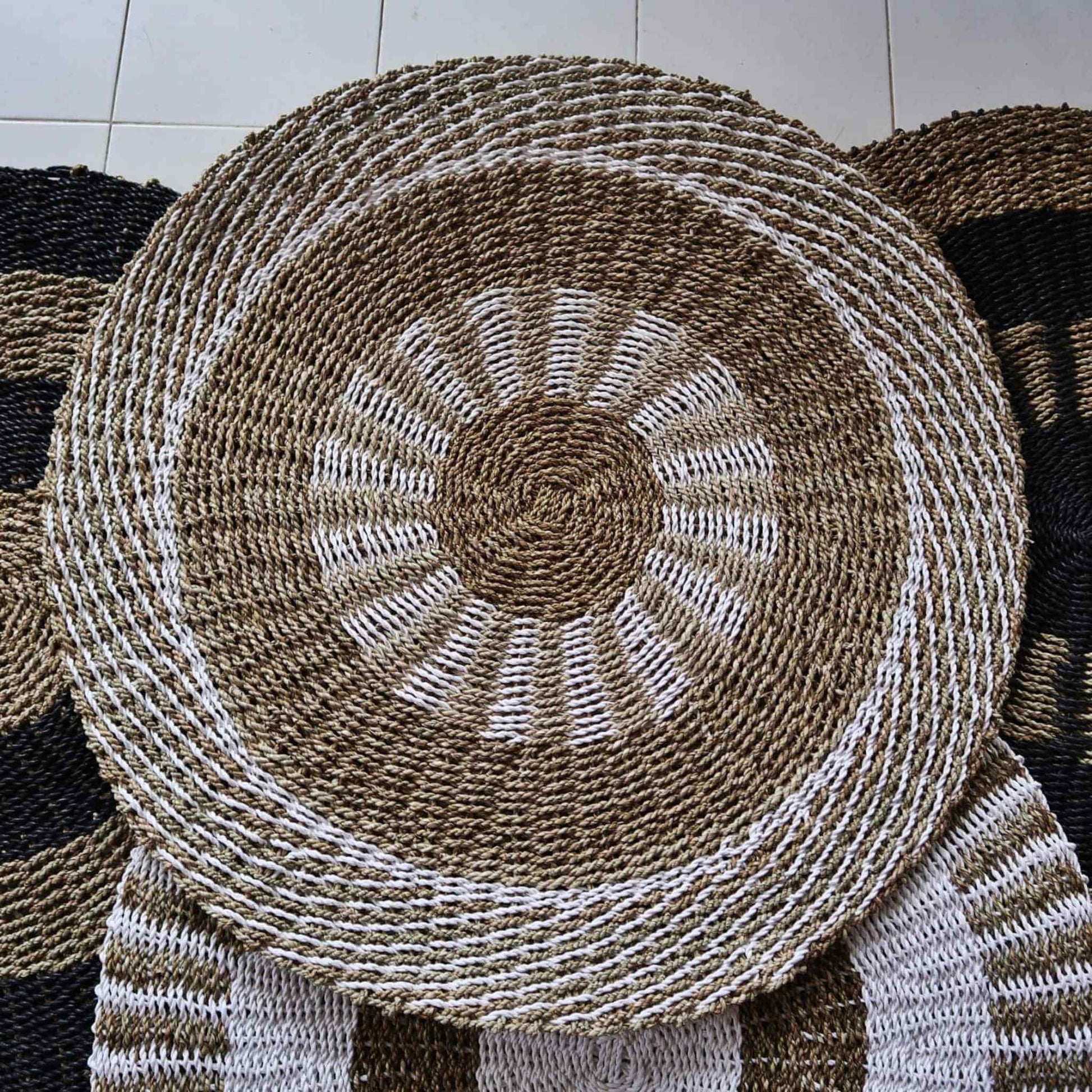 Overhead view of a round seagrass rug with white and tan sunburst design, displayed with other woven rugs.
