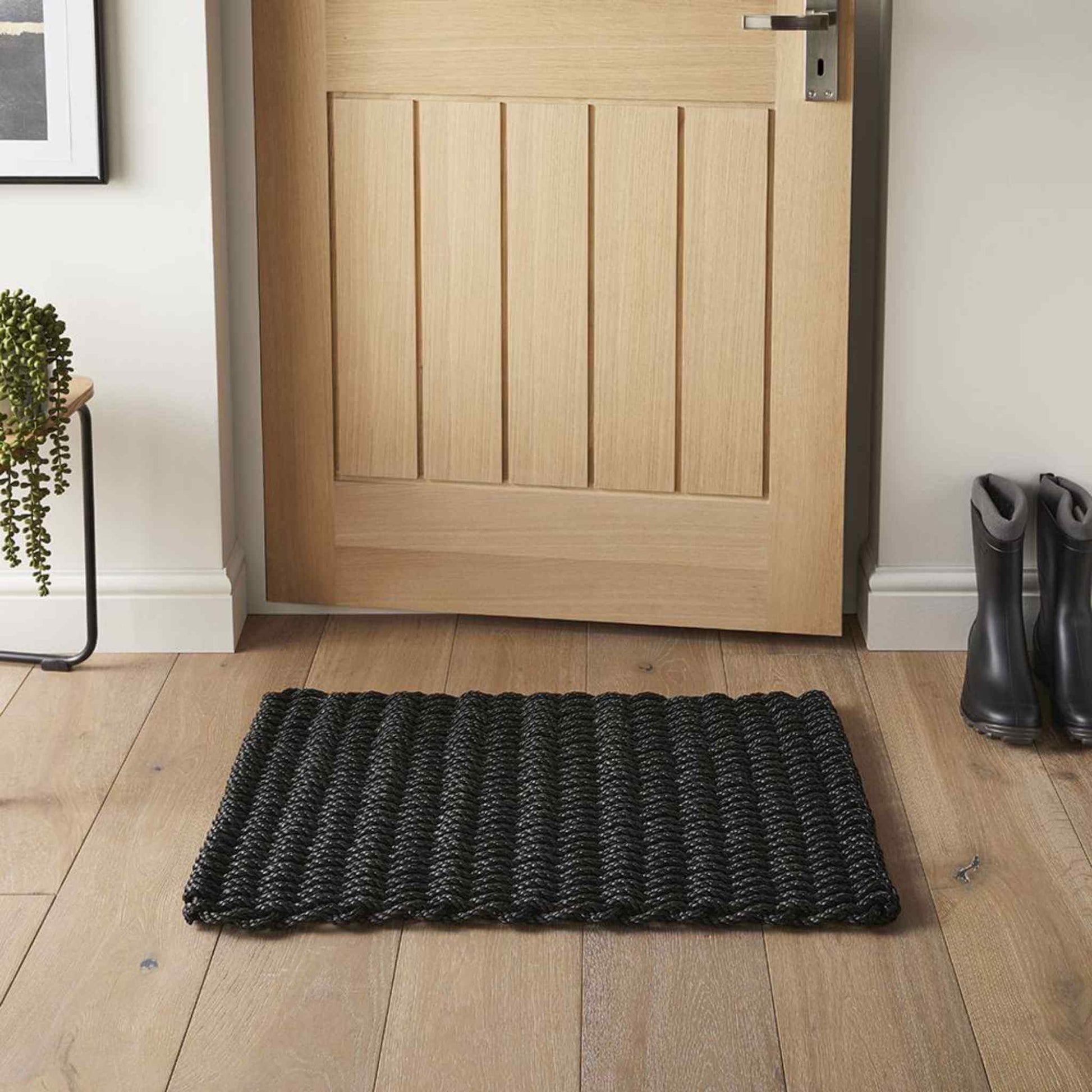 A charcoal braided doormat placed at a front door on a wooden floor beside boots and a plant.