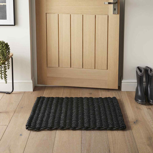 Navy braided polypropylene doormat placed by a wooden door on oak flooring in a hallway setting.