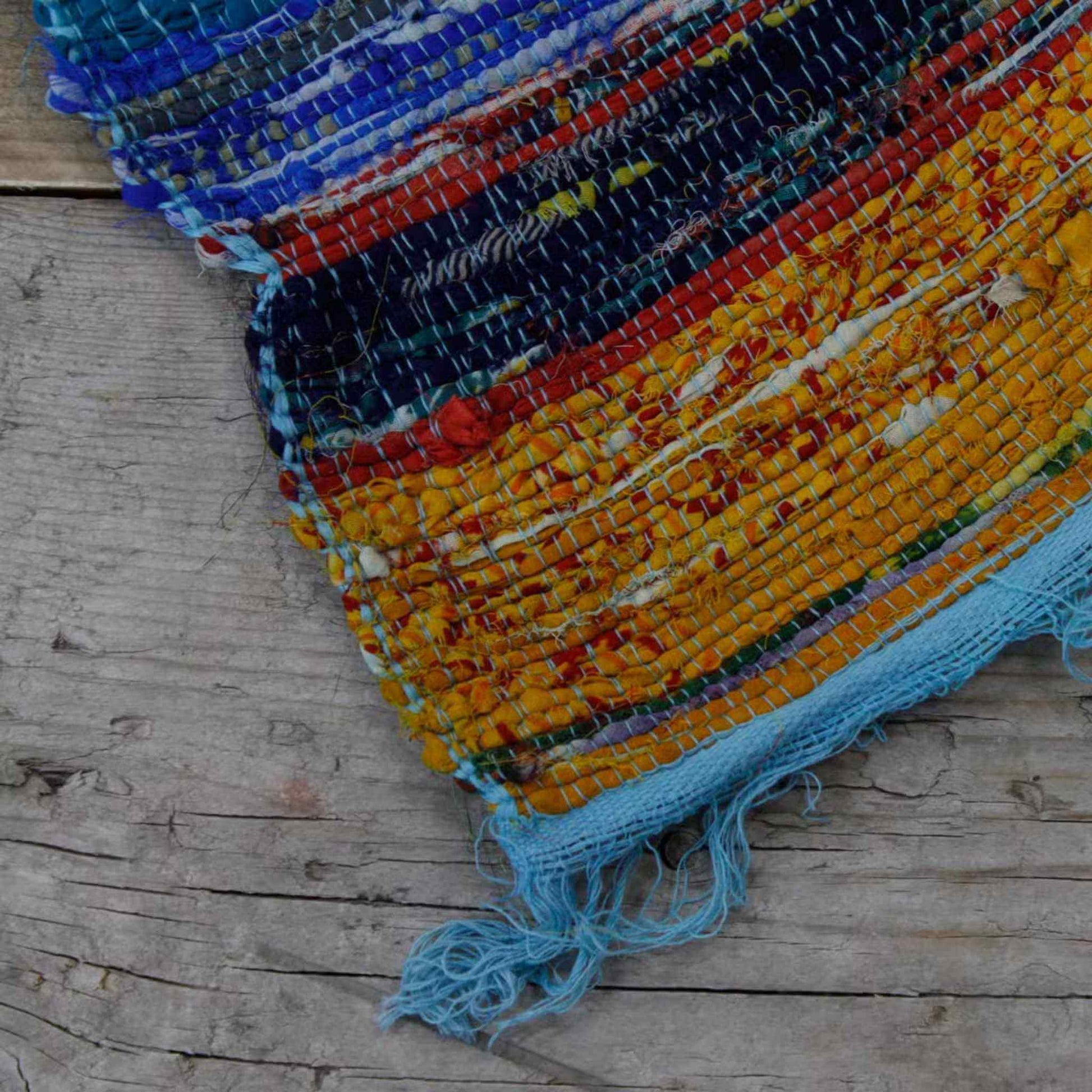Blue fringed corner of an Indian rag rug on a rustic wooden surface, showing handwoven detail.