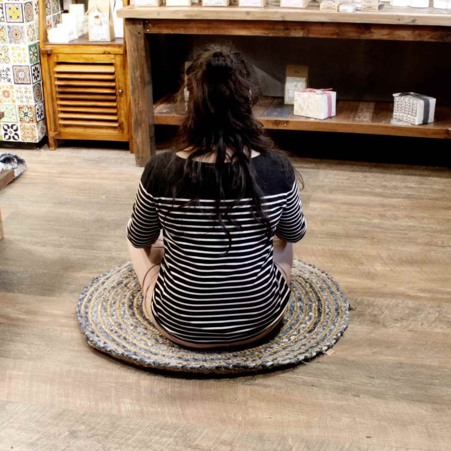 Woman sitting on a 90cm round jute and denim rug in a cosy wooden-floored shop space with rustic shelving.