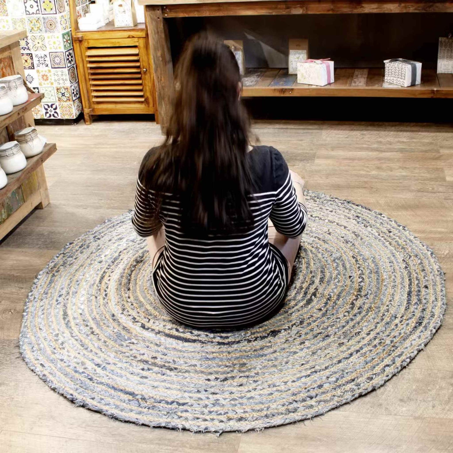 Round jute and recycled denim rug, 150cm wide, in blue and beige tones, displayed on a wooden floor in a shop setting with shelves and homeware products in the background.