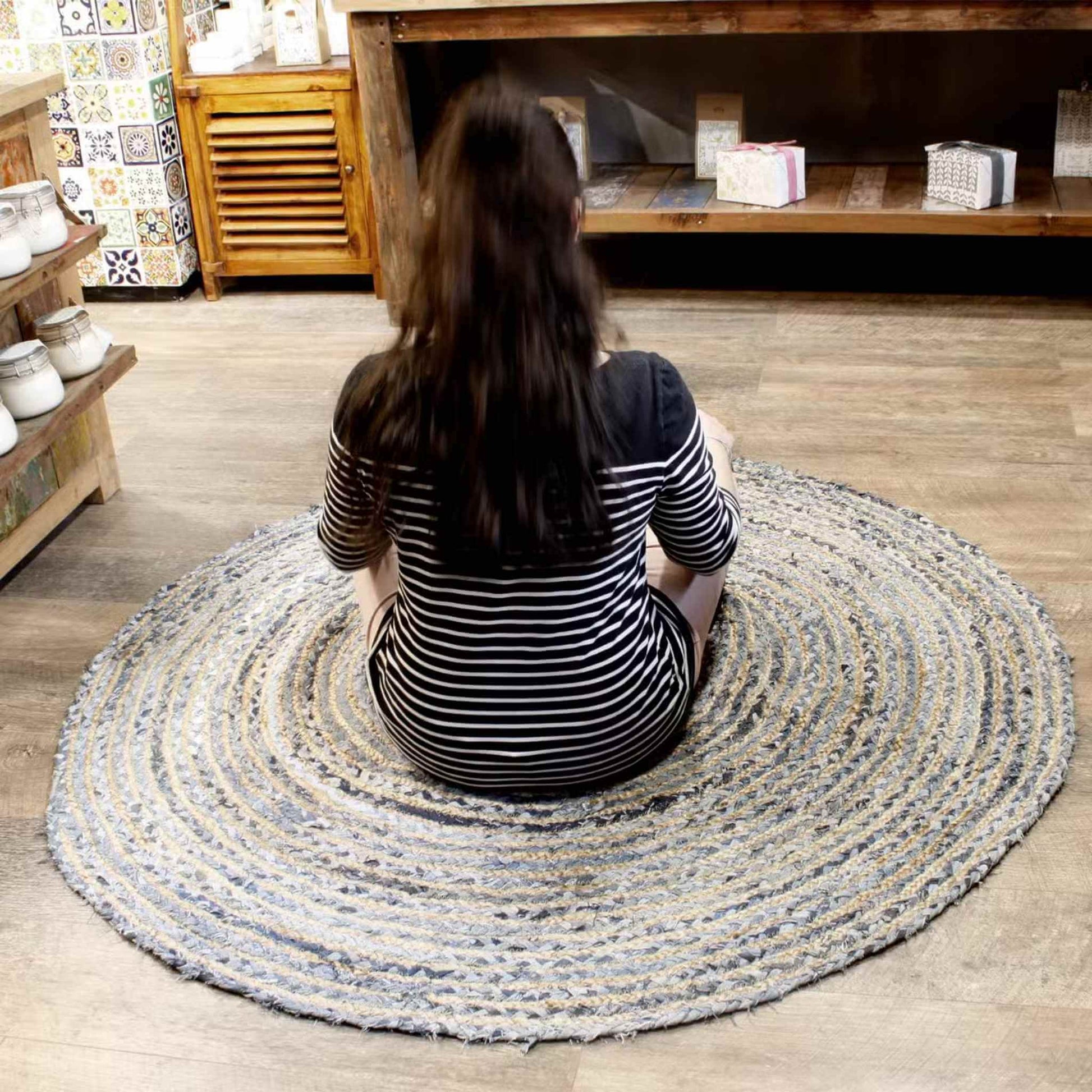 Round jute and recycled denim rug, 150cm wide, in blue and beige tones, displayed on a wooden floor in a shop setting with shelves and homeware products in the background.