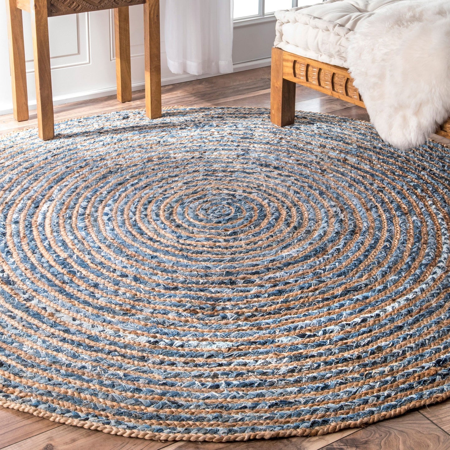 A round jute and recycled denim rug in a light bedroom, set beneath a wooden coffee table and bed.