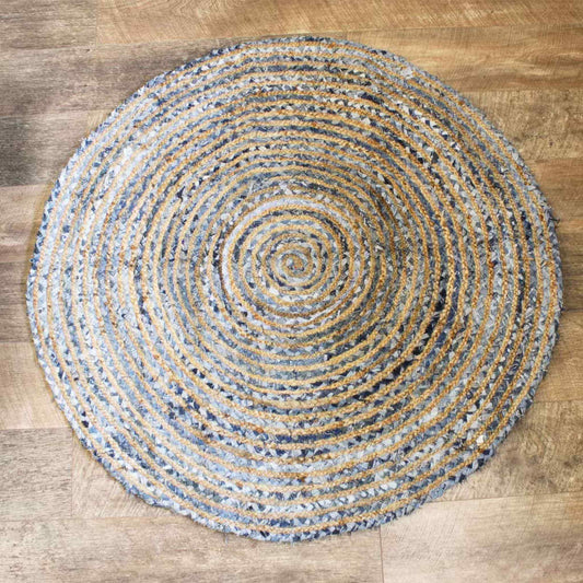 Overhead view of a 120cm round jute and recycled denim rug showing a spiral pattern of blue and beige fibres on a wooden floor.
