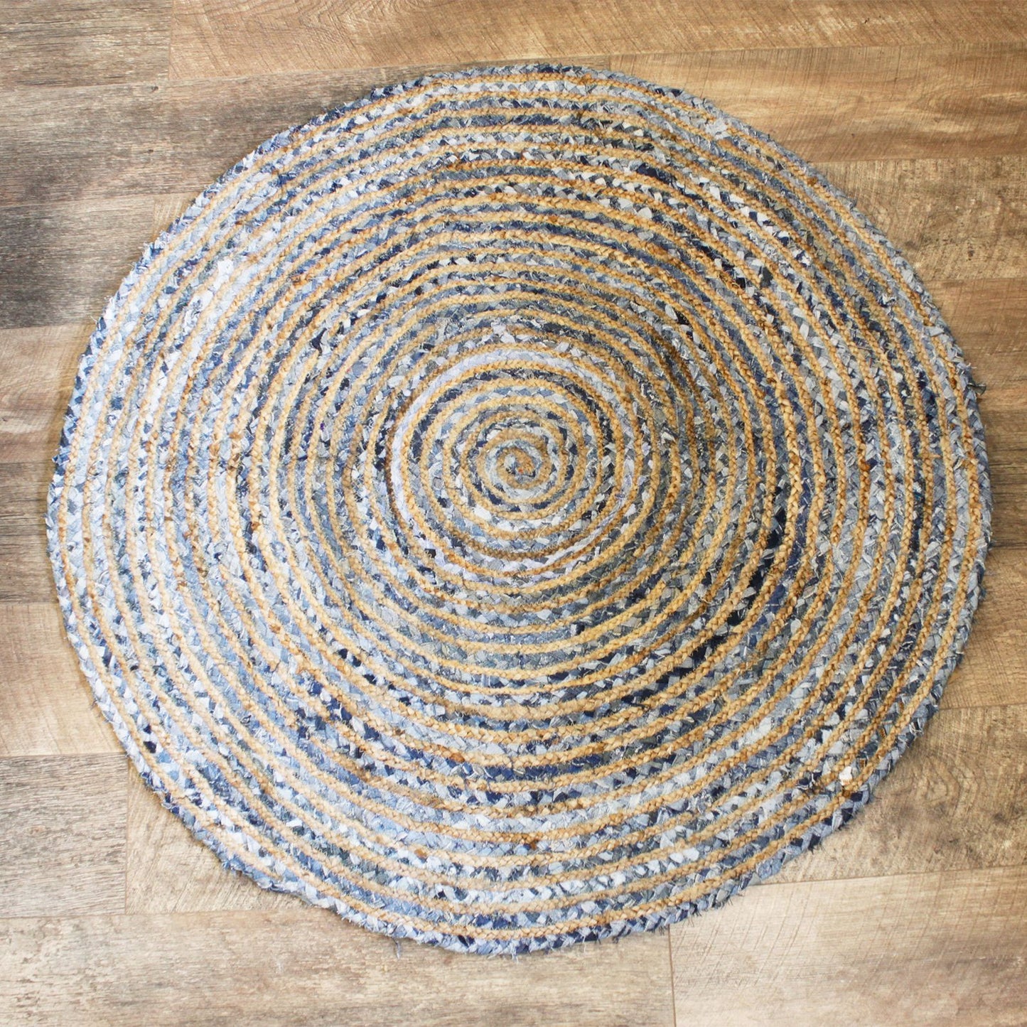 Top-down view of a 120cm round rug made from jute and recycled denim, featuring a coiled pattern in blues, greys, and golds on a wooden floor.
