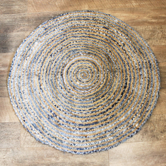 Top-down view of a 120cm round rug made from jute and recycled denim, featuring a coiled pattern in blues, greys, and golds on a wooden floor.