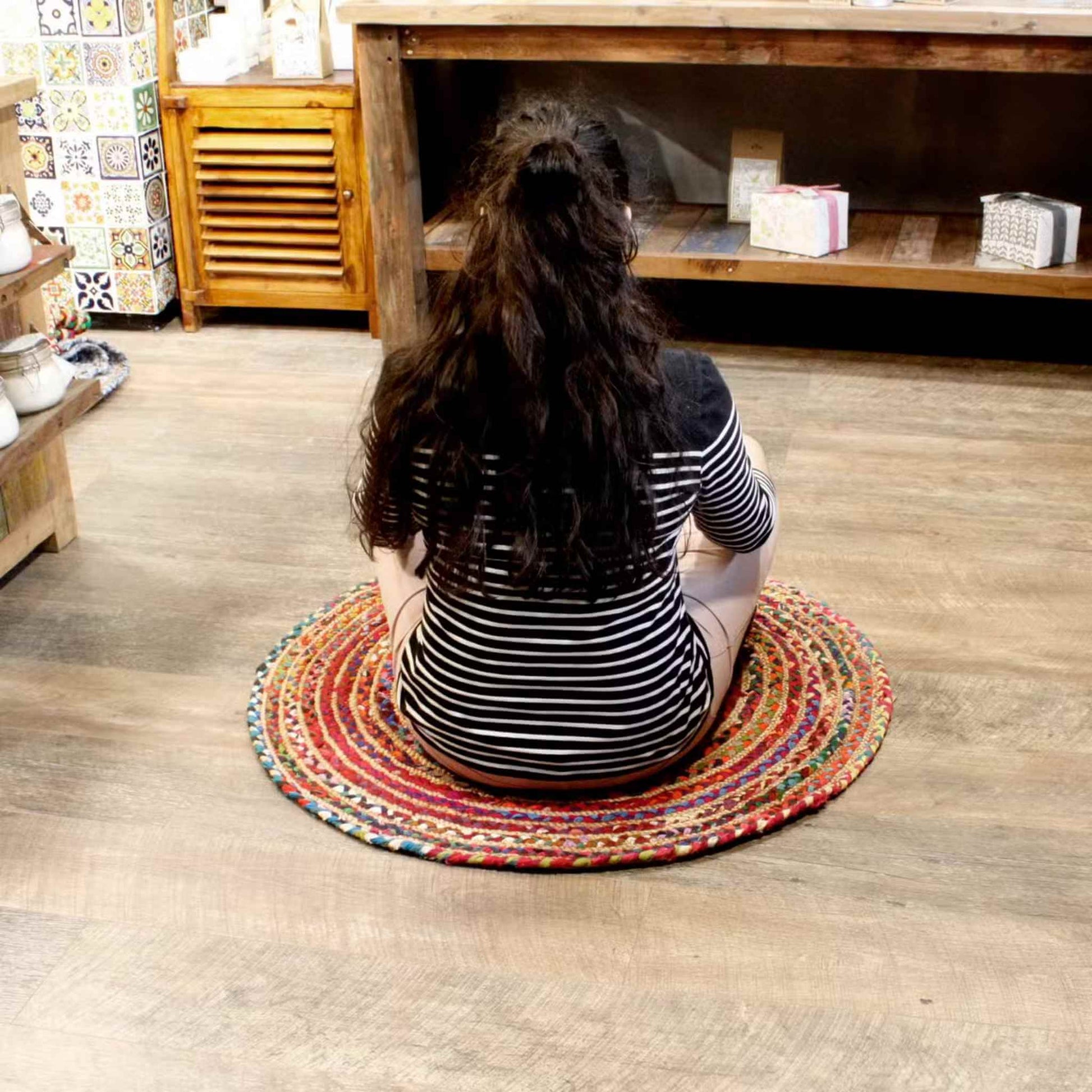 Woman seated cross-legged on a round recycled cotton and jute rug, showing rug scale and cosy indoor styling.