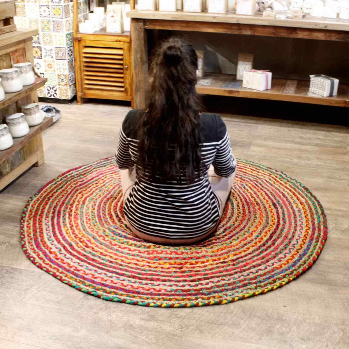 Woman sitting cross‑legged on a large circular jute and cotton rug in a cosy shop setting, demonstrating size and comfort.