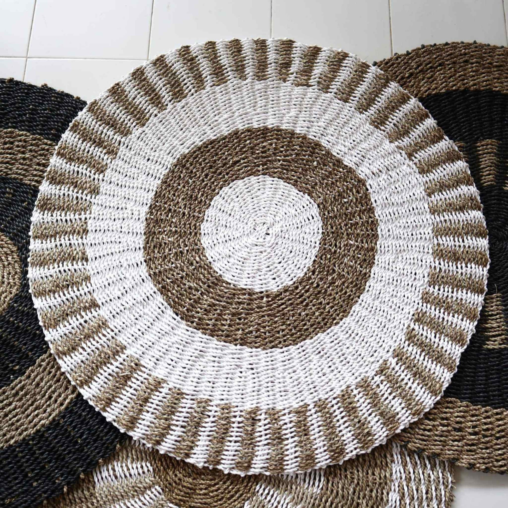 White and tan seagrass rug with radiating sun design displayed with other natural rugs on tiled floor.