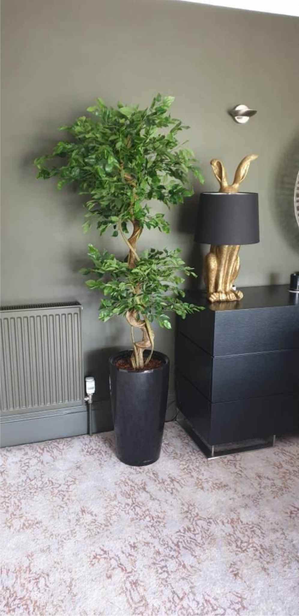 Artificial ficus tree in a tall black pot beside a gold rabbit lamp and a dresser.